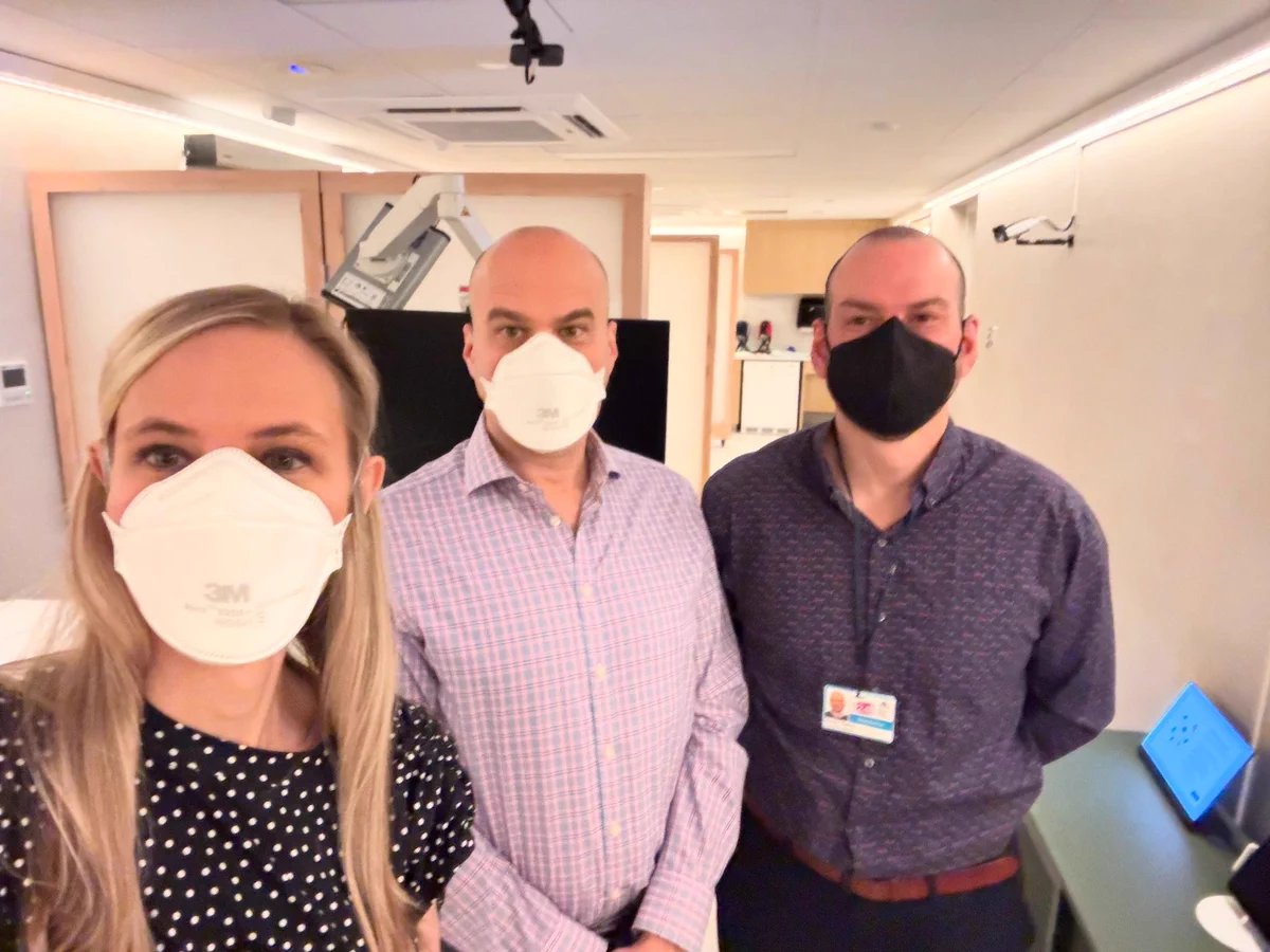 3 people wearing N95 respirators in a hospital clinic. The woman on the left (Amy Proal) is wearing a black T-shirt with white polka dots and has long blonde hair. The man in the middle (Mike VanElzakker) wears a pink and purple checkered collar shirt and has close-cut hair. The man on the right (David Putrino) wears a black respirator and a dark purple long sleeved collar shirt.