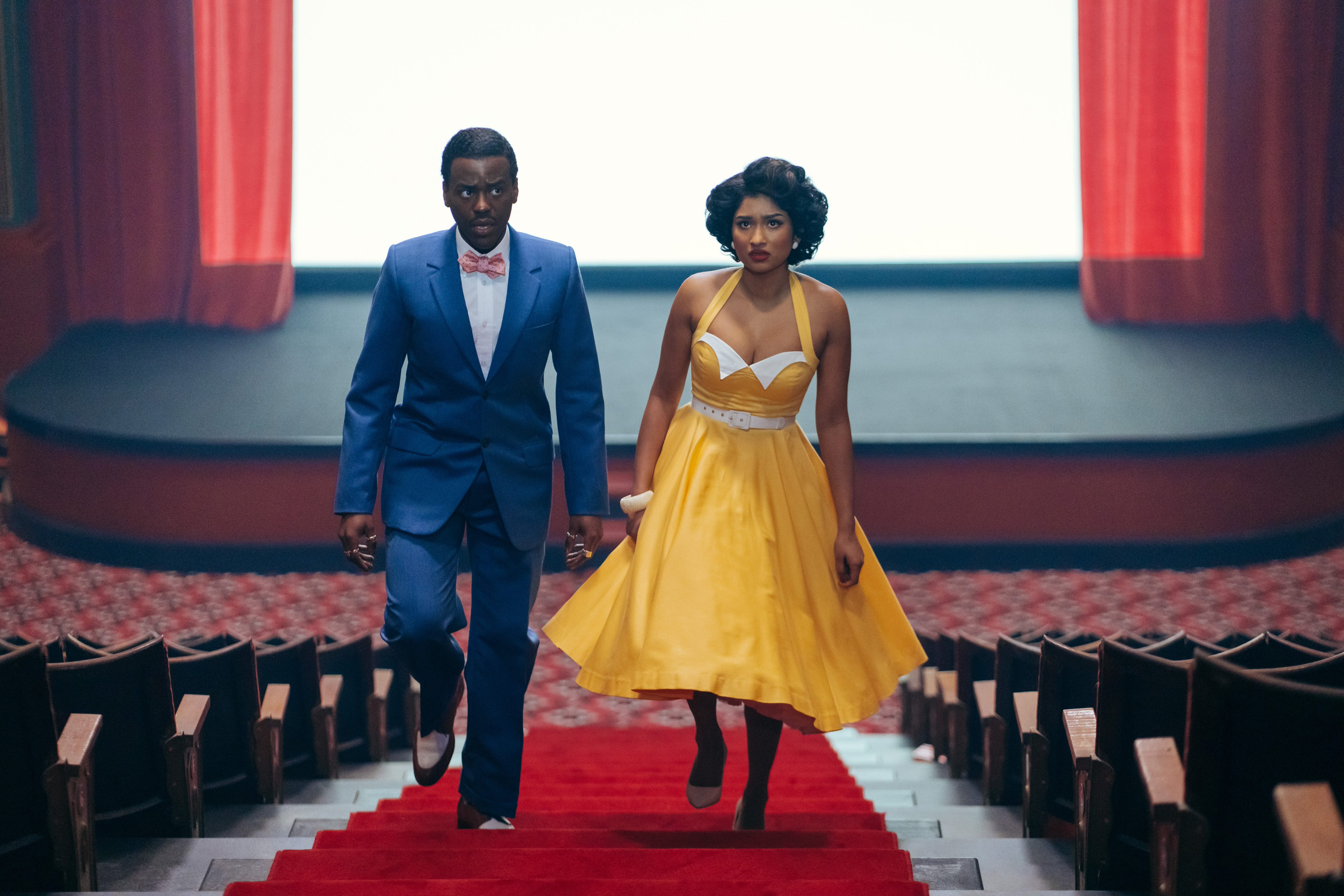 Ncuti Gatwa as the Doctor and Varada Sethu as Belinda Chandra in Doctor Who, dressed in formal attire walking down the aisle of a movie theatre, with a film screen visible in the background.