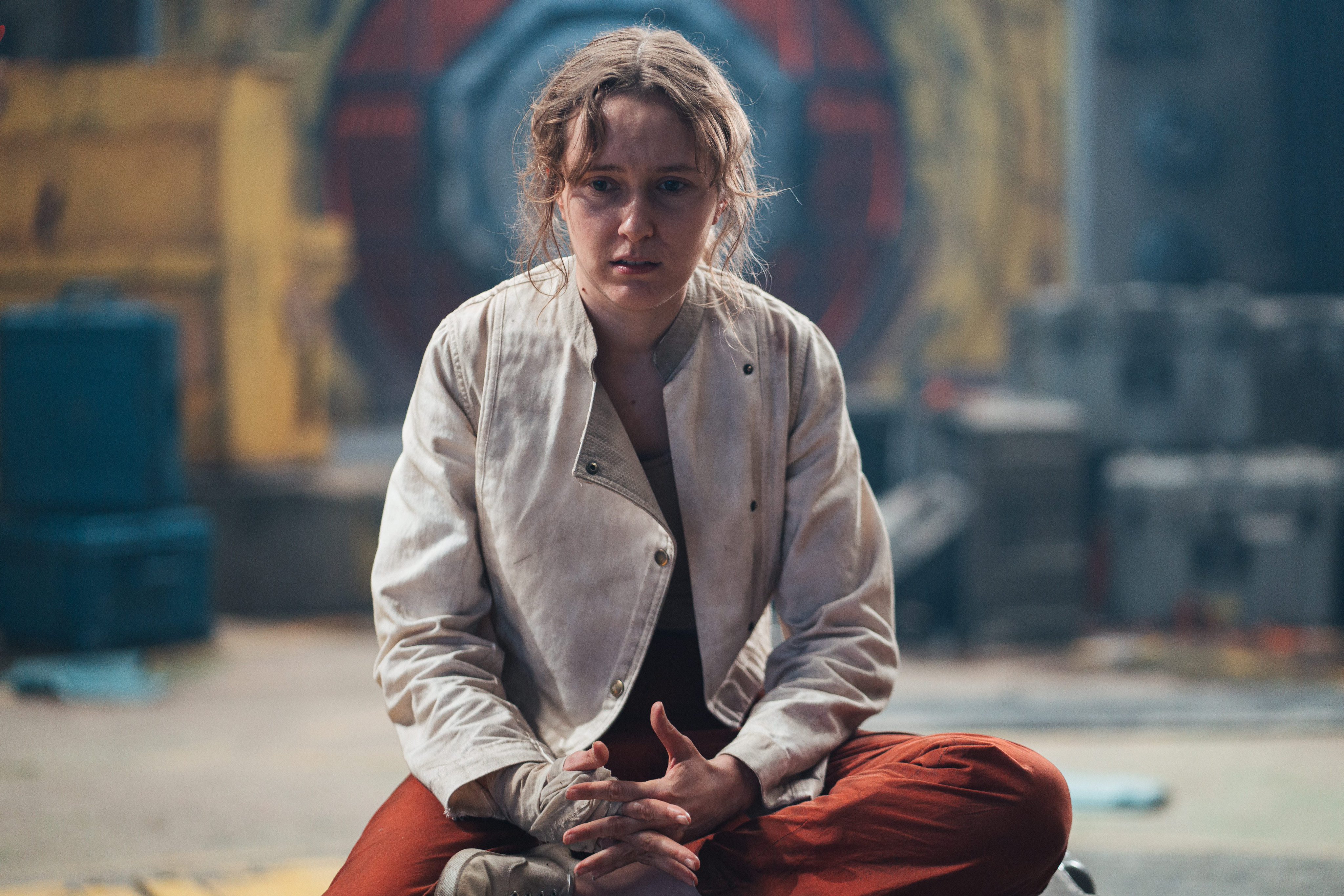 Person sitting on the ground with an intense expression, dressed in a light jacket and red pants, set against a background featuring a large, elaborate symbol.
