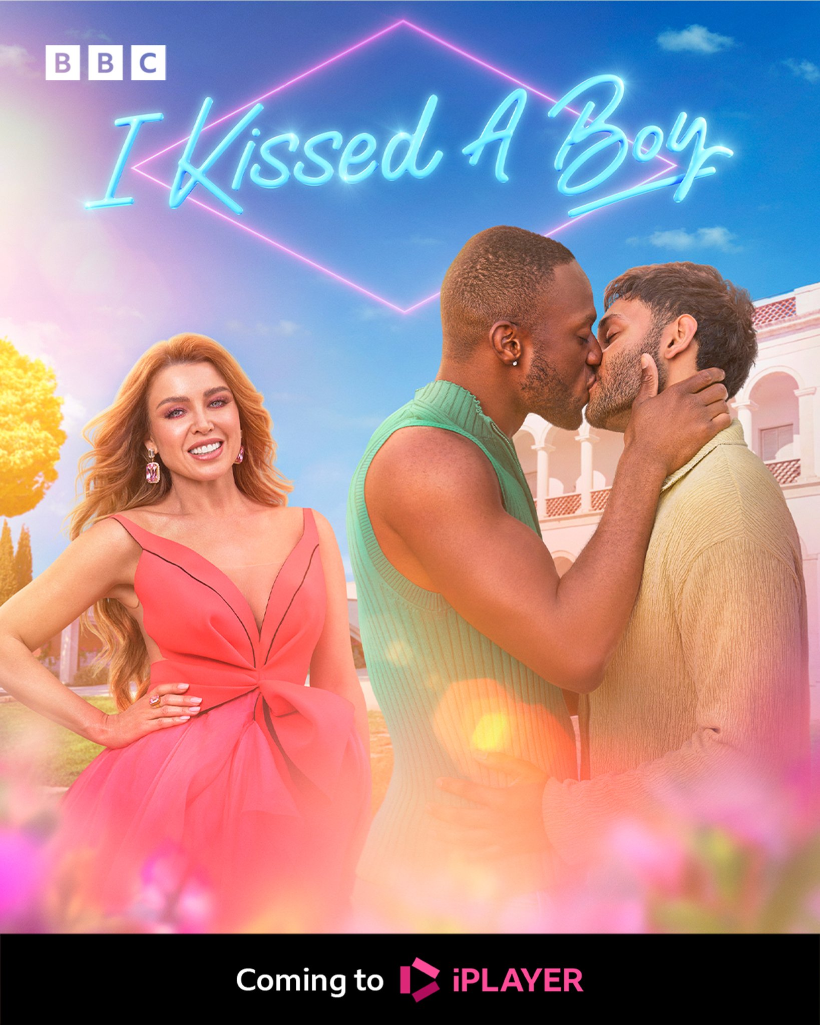 Promotional image for I Kissed A Boy. Dannii Minogue in a bright pink dress smiling at the camera, standing beside two boys sharing a kiss. Behind them is the sunny Masseria under a clear blue sky. At the top, neon text reads "I Kissed A Boy". At the bottom, the text says "Coming to iPlayer"