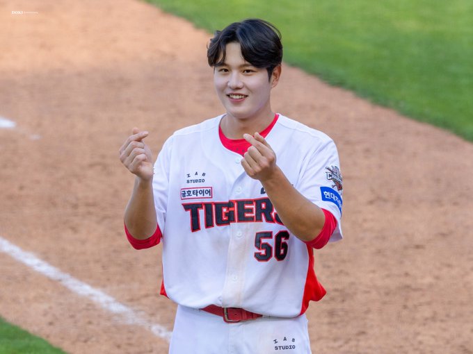 The image shows a baseball player from the Kia Tigers team, wearing a white and red uniform with the number 56. The player is on a baseball field, with the green grass and dirt of the diamond visible in the background. He is making a playful gesture with his hands, smiling, which suggests a light-hearted moment during or after a game. The uniform has the team name 'Tigers' and the player's name '오선우' (Oh Seon-woo) written on it. The post text includes emojis 🫰😁🫰, indicating a cheerful and playful mood. The hashtags #기아타이거즈 (Kia Tigers) and #오선우 (Oh Seon-woo) confirm the player's identity and team affiliation.