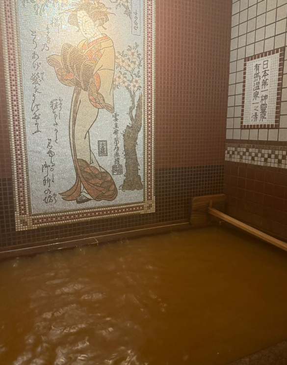 壁のモザイク壁画には伝統的な着物姿の女性が描かれており、日本語の書で飾られています。お湯はこげ茶色で、ミネラルたっぷりの天然温泉なのか、和風の風情が漂っています。こんな温泉に入ったことはありませんか?🥰