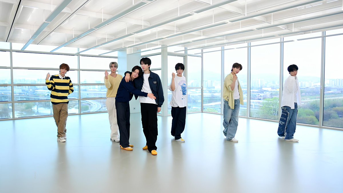 Seven members of a K-pop boy group pose in a bright, modern room with large floor-to-ceiling windows overlooking a city skyline.