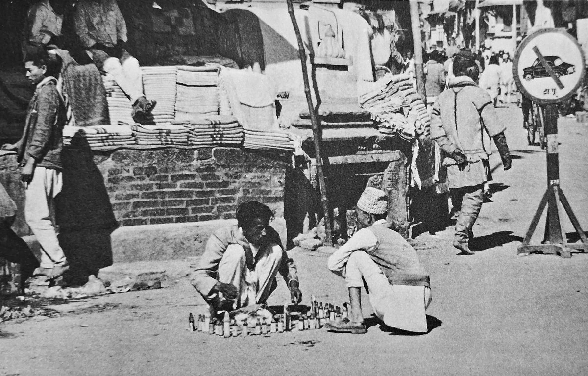 In the 1970s. A 'No Cars' sign is visible on the right. The location is unidentified. In the foreground, men are seen buying and selling local medicines.