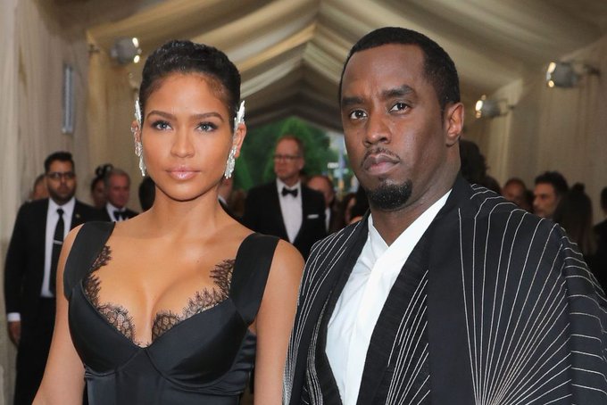 La imagen muestra a dos individuos, presumiblemente Diddy y Cassie Ventura, vestidos con atuendos formales en lo que parece ser un evento de alto perfil. La mujer lleva un vestido negro elegante con detalles de encaje, mientras que el hombre lleva un traje blanco y negro con un diseño único. El escenario es un lugar interior glamoroso con otros asistentes vestidos formalmente en el fondo, lo que sugiere una alfombra roja o un evento similar. Dado el contexto de la publicación, es probable que esta imagen se use para ilustrar las personalidades públicas de Diddy y Cassie Ventura en medio de las graves acusaciones discutidas en el juicio, incluidas las drogas, el chantaje y los rituales sexuales prolongados.