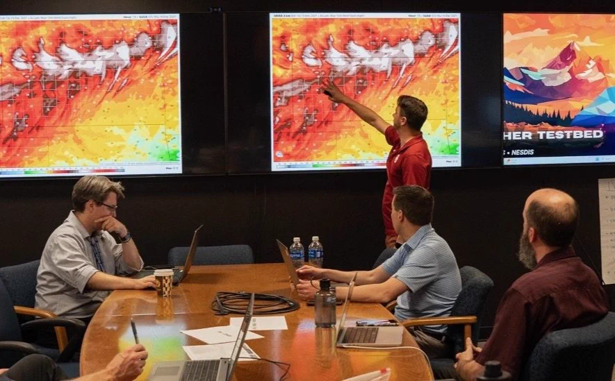 An individual presenting weather data on large screens in a meeting room with several experts focused on the discussion. Photo credit: NOAA Research.