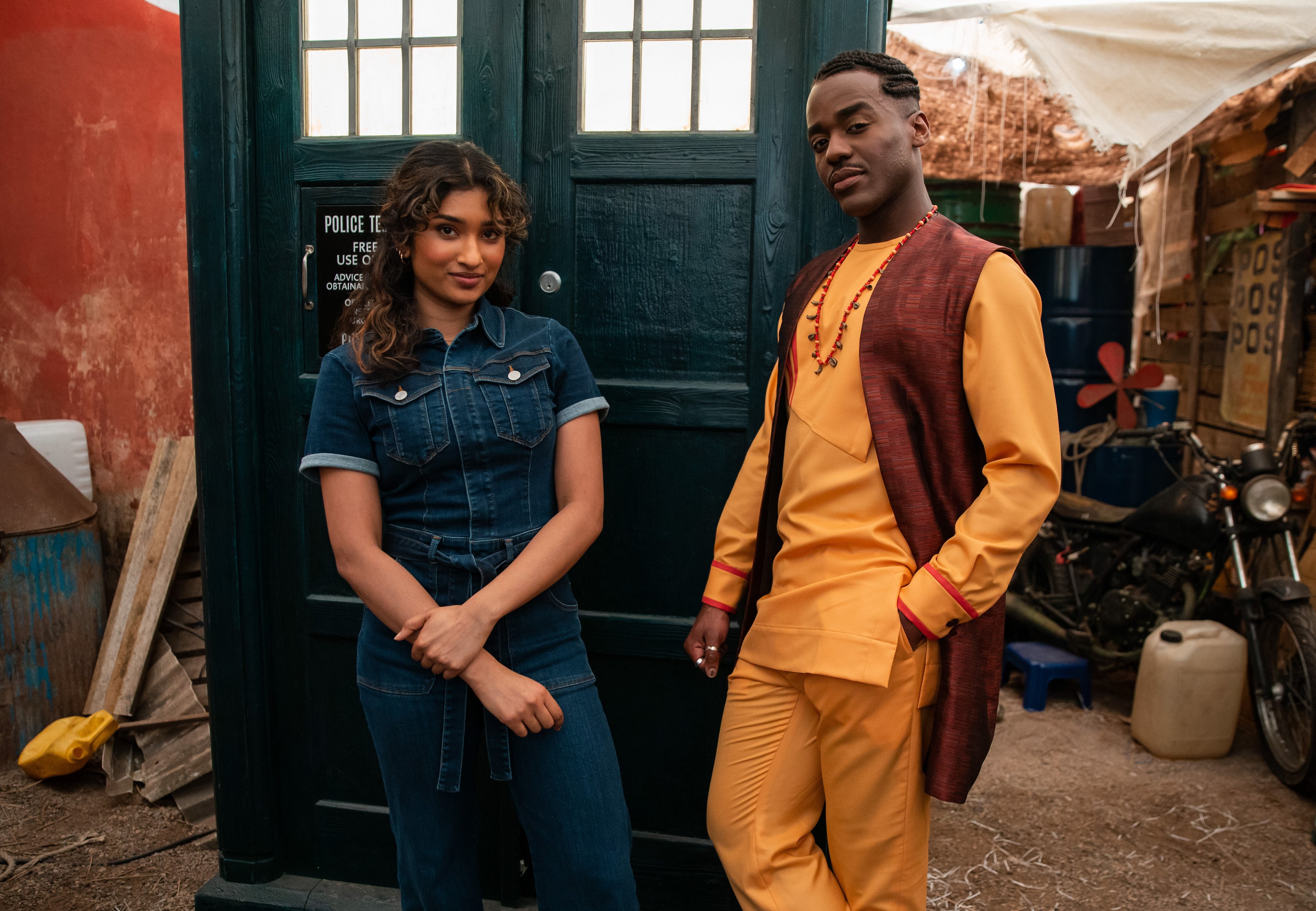 Two individuals Ncuti Gatwa as The Doctor and Varada Sethu Belinda Chandra standing in front of the TARDIS from Doctor Who, dressed in contemporary outfits.