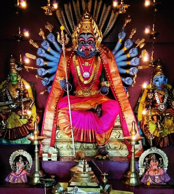 The image depicts a vibrant and detailed statue of Pratyangira Devi, a fierce Hindu goddess known for her protective and purifying powers. She is shown with multiple arms, each holding various symbolic items, and is adorned with traditional jewelry and vibrant clothing. Flanking her are two smaller statues, likely representing other deities or attendants. The setting is a richly decorated altar, illuminated by numerous lamps, enhancing the divine and mystical atmosphere. According to the post, Pratyangira Devi is also referred to as Gayatri, embodying the hidden fire within the Gayatri mantra, a powerful cosmic force that dissolves darkness and protects dharma. This depiction aligns with the Tantric tradition where Gayatri is seen as both a source of light and a weapon against negative forces.