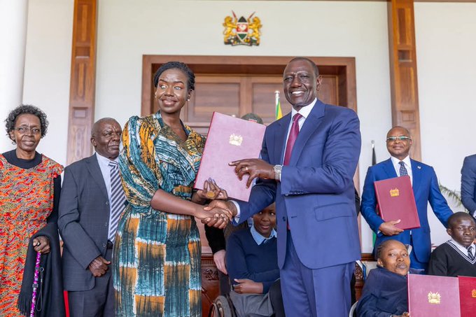 Senator Crystal Asige stands beside the President of Kenya  shaking hands  holding the Act. She is wearing an orange, white, and blue tai and dai kitenge dress, with her afro hair styled and tied back. The President is dressed in a navy blue suit, white shirt, and red tie.