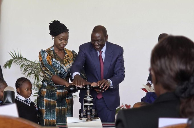 Senator Crystal Asige stands beside the President of Kenya as they jointly press a metallic seal onto the Persons with Disabilities Bill, enacting it into law. She is wearing an orange, white, and blue tai and dai kitenge dress, with her afro hair styled and tied back. The President is dressed in a navy blue suit, white shirt, and red tie.