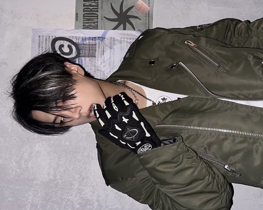 Rocking an effortlessly cool vibe, this stylish commands attention in a sleek olive-green bomber jacket, layered over a crisp white tee and adorned with a subtle pendant necklace. The black gloves with bold, edgy designs add a touch of mystery and rebellion.