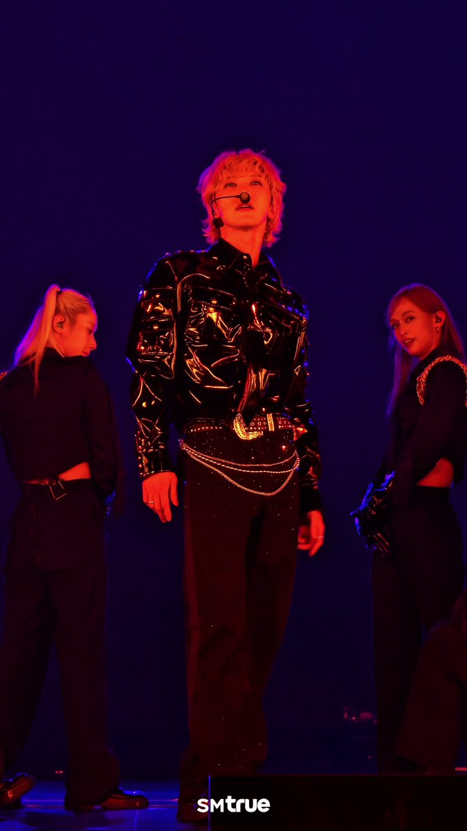A performer with blonde hair, wearing a shiny black jacket and headpiece, stands center stage illuminated by blue lighting, flanked by two backup dancers; the watermark 'smtrue' appears at the bottom.