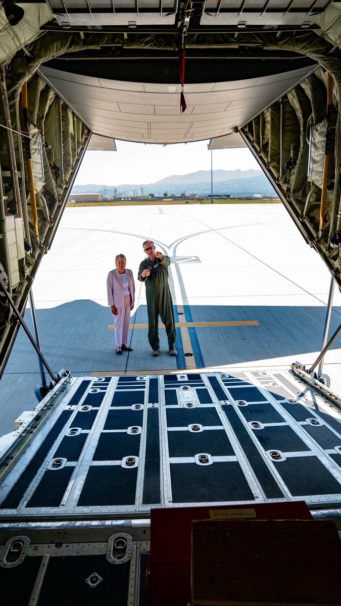 The image shows the interior view from the back of a large aircraft, looking out onto a tarmac. Two people are standing outside on the tarmac, one in a light-colored outfit and the other in a darker uniform. The aircraft door is open, and the tarmac, mountains, and some buildings are visible in the background. There is some text present on the aircraft.