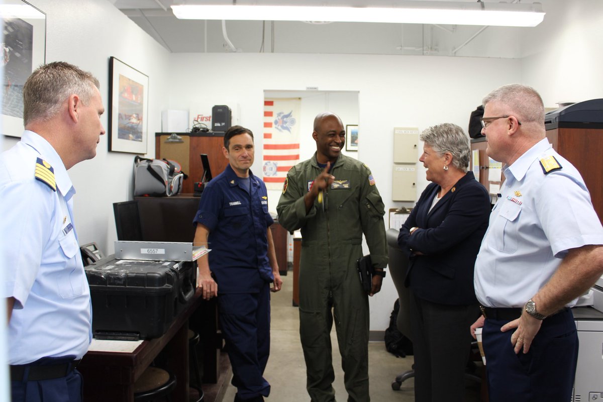 The image shows five people in a small office setting, engaged in conversation. They are wearing uniforms and appear to be in a professional or military environment. There are various objects in the room, including a black case, wall decorations, and a computer monitor.