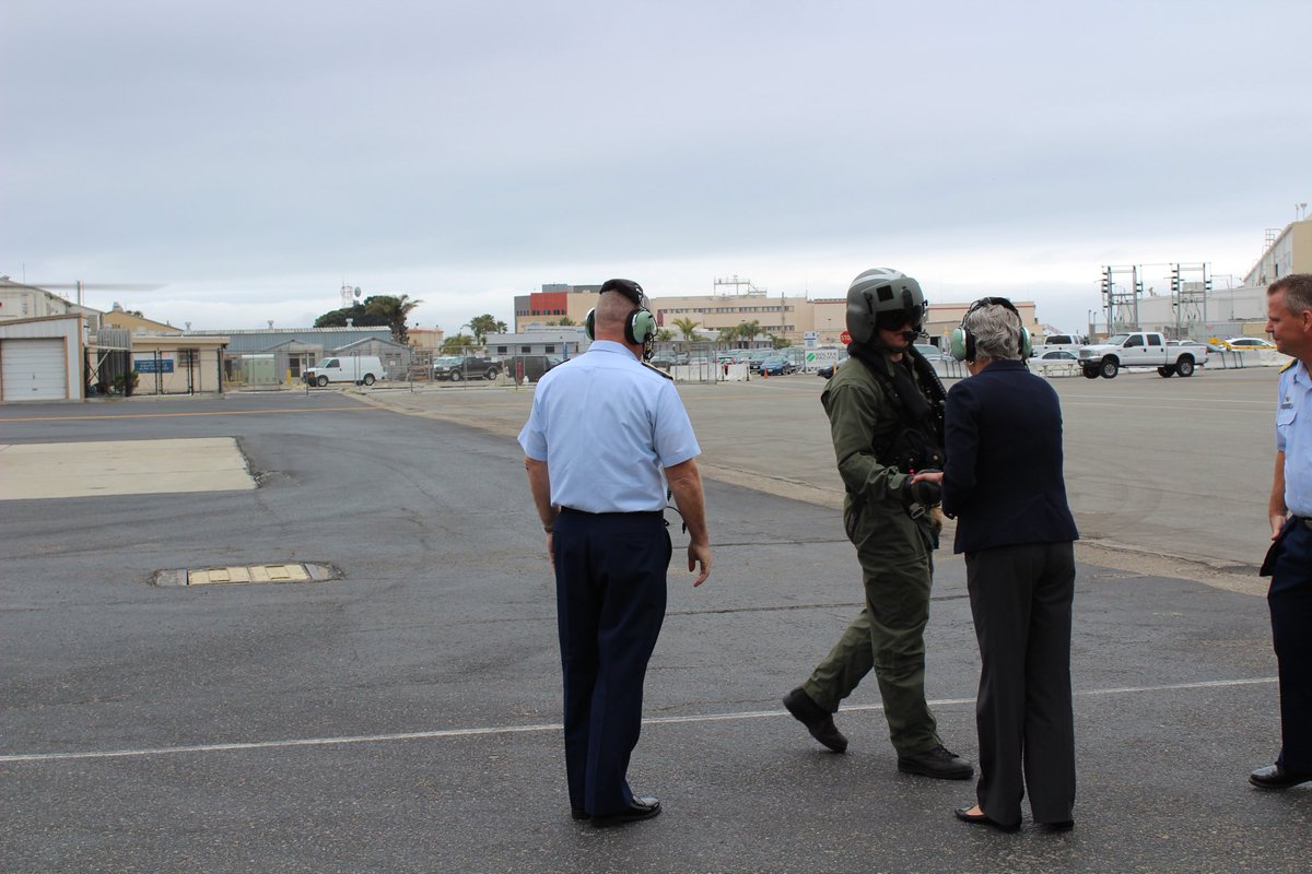 The image shows several people on an airfield. One person is in a flight suit and helmet, shaking hands with another person wearing a headset. There are buildings, vehicles, and a cloudy sky in the background. There is text on some signs.