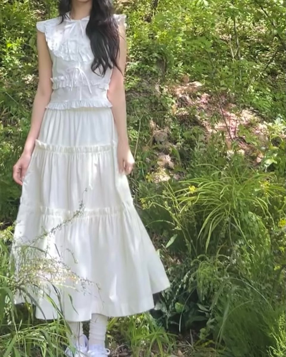 jiaheon's tweet photo. A serene spring park with tall green trees, soft grass, and wildflowers. A woman in a flowing white dress walks through the scene, sunlight filtering through leaves, with gentle wind lifting her hair and skirt. https://t.co/sMCRy1vV2K