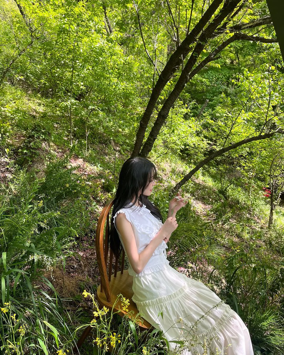 jiaheon's tweet photo. A serene spring park with tall green trees, soft grass, and wildflowers. A woman in a flowing white dress walks through the scene, sunlight filtering through leaves, with gentle wind lifting her hair and skirt. https://t.co/sMCRy1vV2K