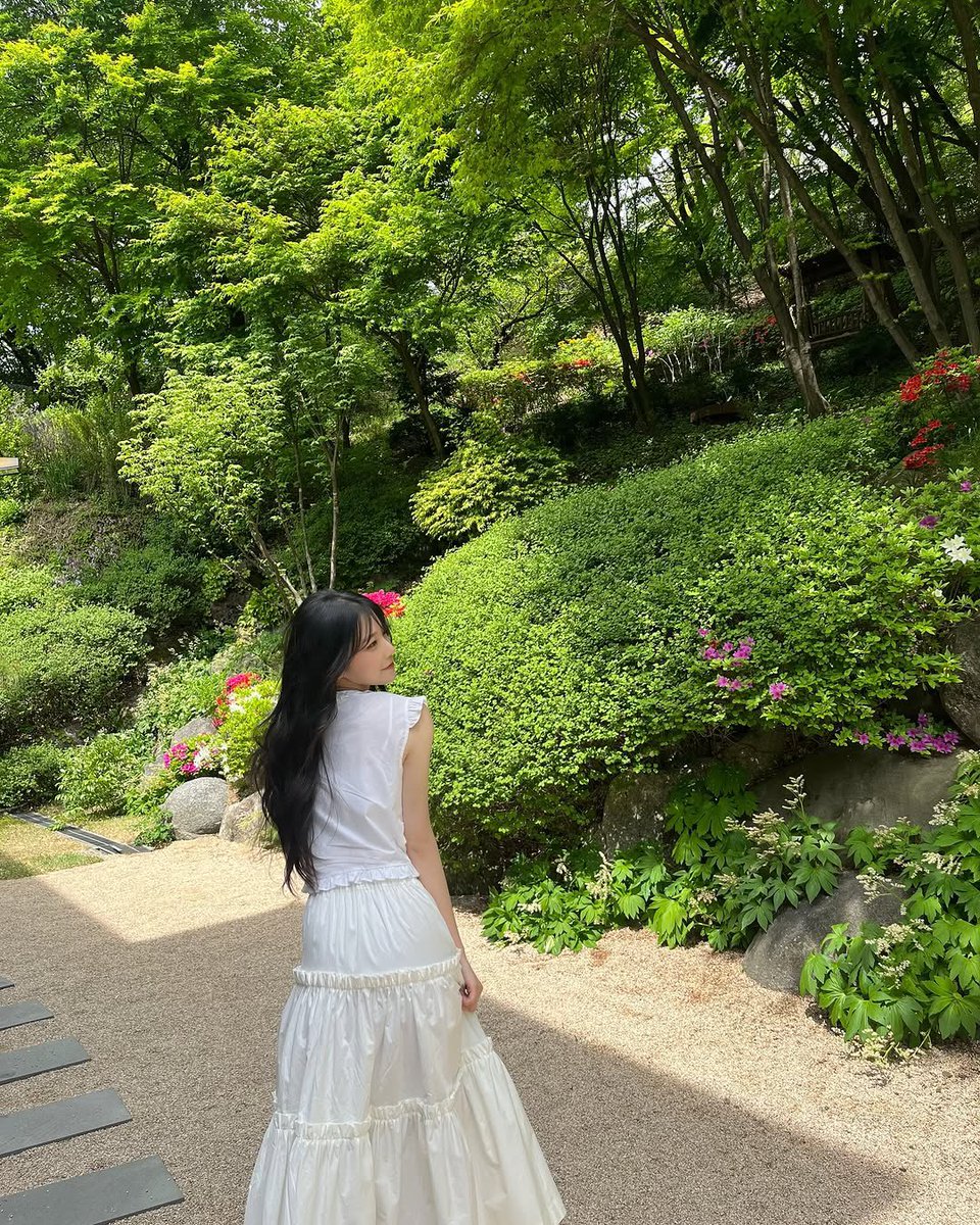 jiaheon's tweet photo. A serene spring park with tall green trees, soft grass, and wildflowers. A woman in a flowing white dress walks through the scene, sunlight filtering through leaves, with gentle wind lifting her hair and skirt. https://t.co/sMCRy1vV2K