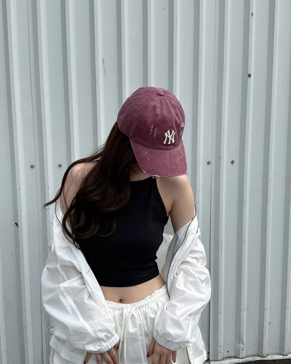 A soft gaze beneath a worn maroon cap, with wind-tousled waves cascading down like ink. Draped in a cloud-white jacket over jet-black calm, she stands against the silver wall — quiet, effortless, and entirely her own.