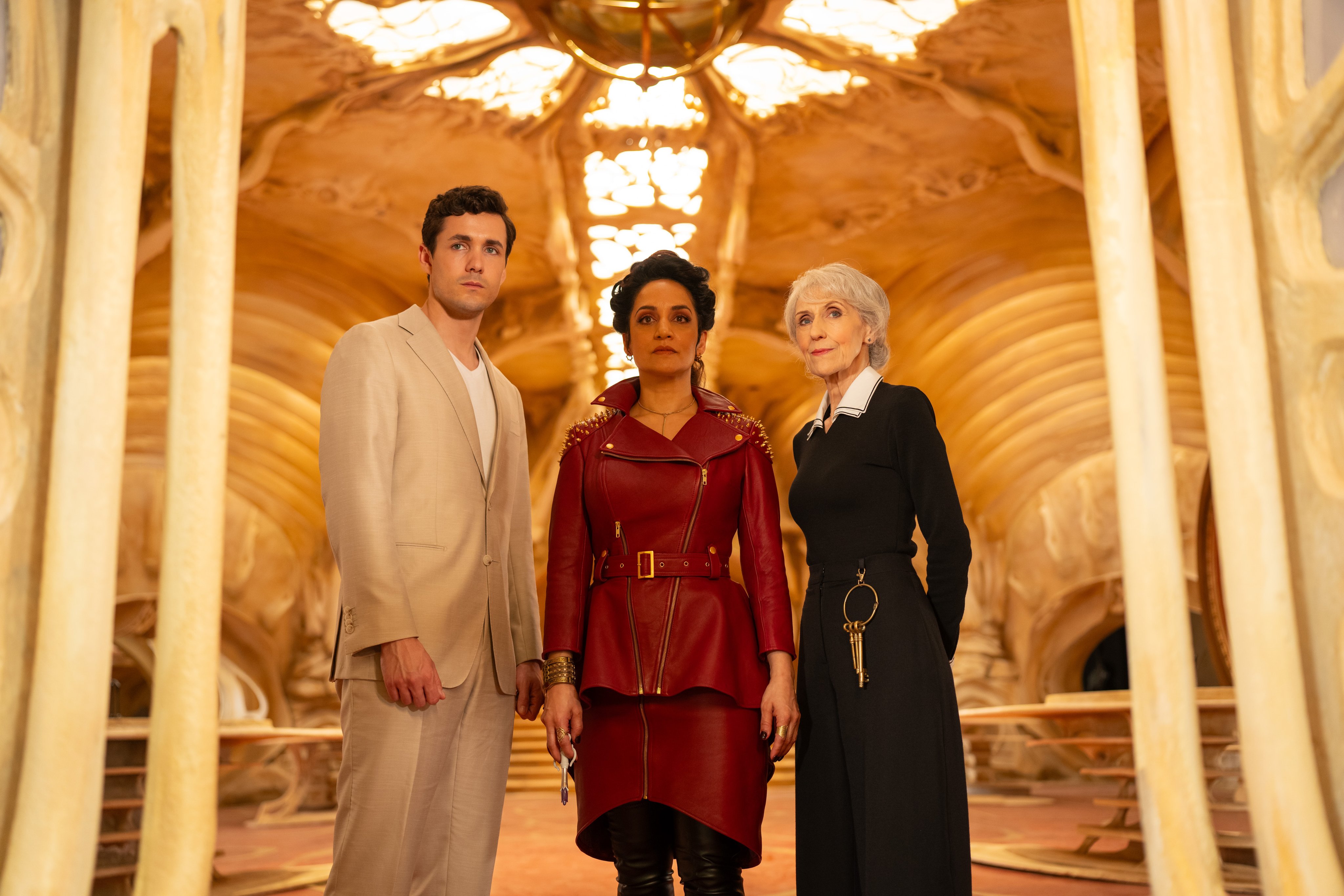 Jonah Hauer-King, Archie Panjabi and Anita Dobson standing side by side in an ornately decorated room with golden hues and elaborate architectural details.. 