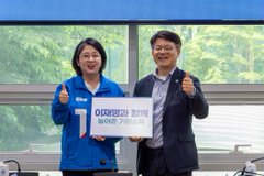 선거운동복을 착용한 용혜인 선대위원장과 검은 정장을 입은 김철호 기본소득위원회 위원장이 ‘이재명과 함께 농어촌 기본소득’이라는 피켓을 들고 웃으며 엄지손가락을 들어 보이고 있다. 창밖에는 초록 나무들이 보인다.