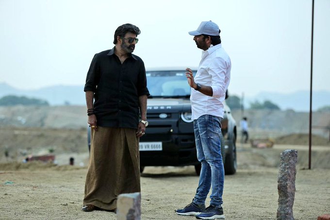 The image depicts two men standing in an outdoor setting, likely a film set, with a rugged, earthy background. One man is dressed in a black shirt and traditional dhoti, while the other wears a white shirt, blue jeans, and a cap, suggesting a casual yet professional interaction. Behind them is a black SUV, adding to the action-oriented ambiance of the scene. Given the context from the post text, this image is related to the upcoming film where Nandamuri Balakrishna, known as #Balayya, plays a dual role under the direction of Gopichand Malineni. The post hints at a film filled with action and emotions, with Balakrishna sporting a mass rugged look, indicating this could be a behind-the-scenes shot from the movie production.