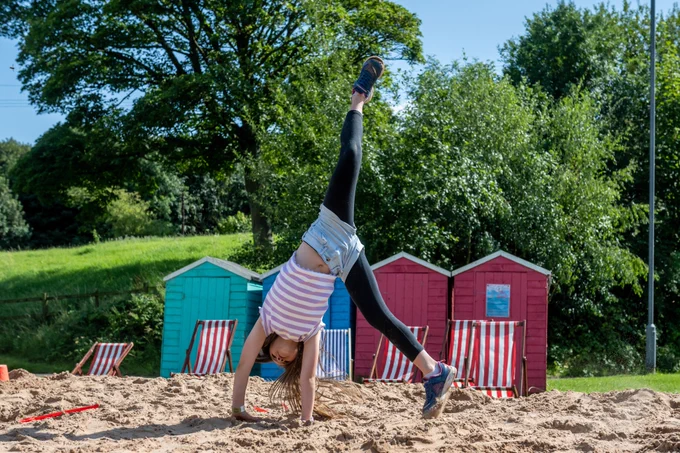 Child doing a cartwheel on the sand in front of five deck chairs and four beach huts 