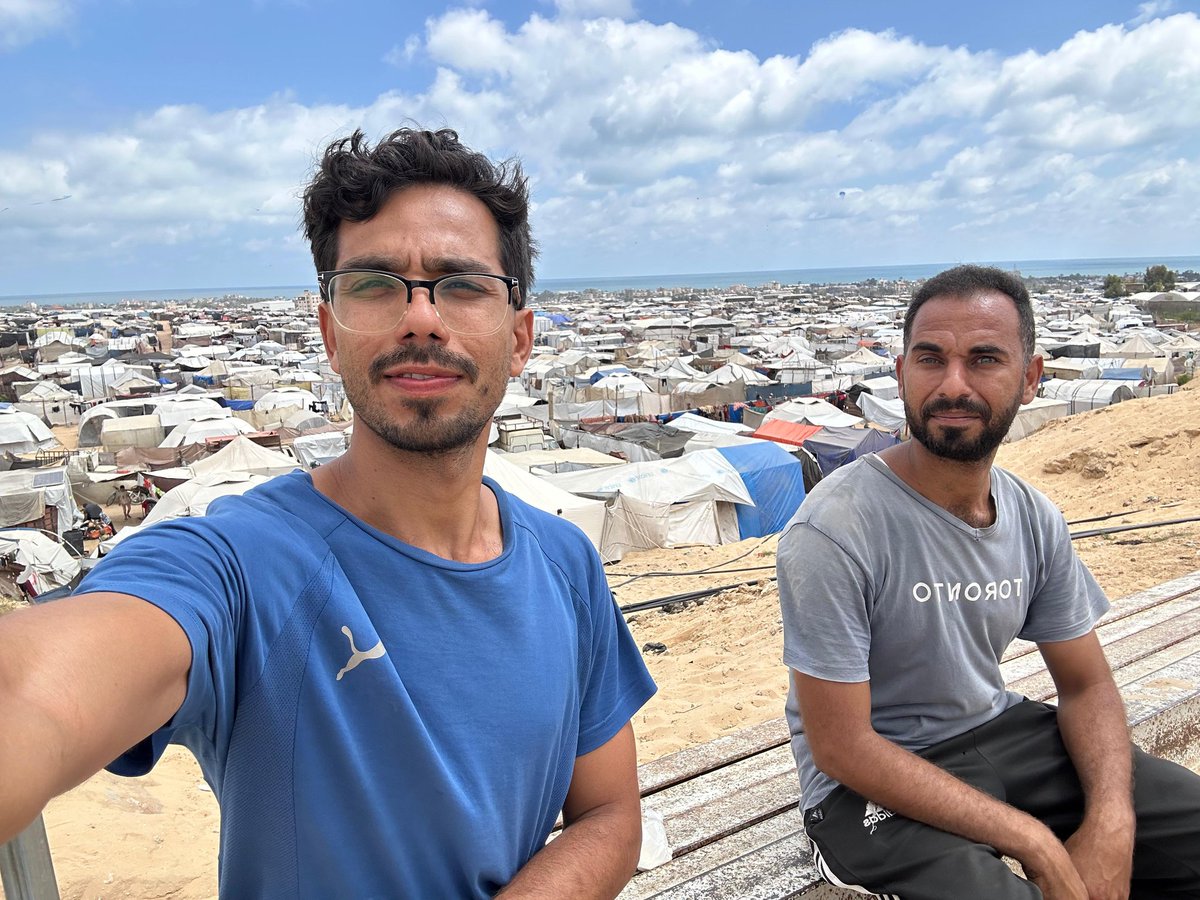 Me, my brother, and my friend Ibrahim stand in the al-Mawasi tents in Khan Younis, after the war displaced us and destroyed our homes. Rubble surrounds us, but our pain is not just from the destruction—it’s the hunger slowly killing our children. There’s no electricity, no clean
