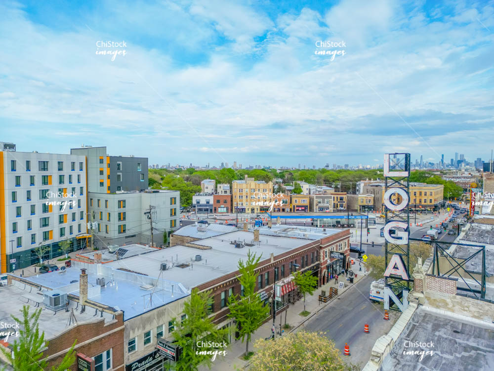 Logan Theatre Sign on Milwaukee Ave with CTA Station in #LoganSquare. High-resolution photo of the neon Logan Theatre sign above Milwaukee Avenue. Shows the Logan Square #CTA Blue Line entrance across the street. Brick rooftops and green trees frame the scene.