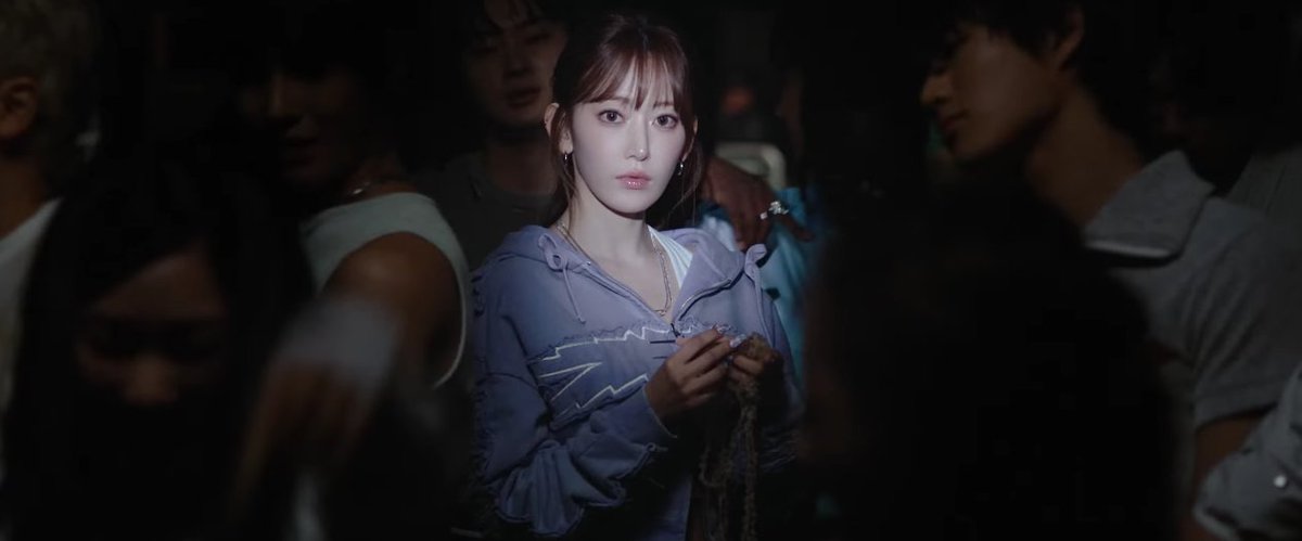 A young woman with bangs stands out in a crowd, wearing a light blue top with ruffled details and a necklace.