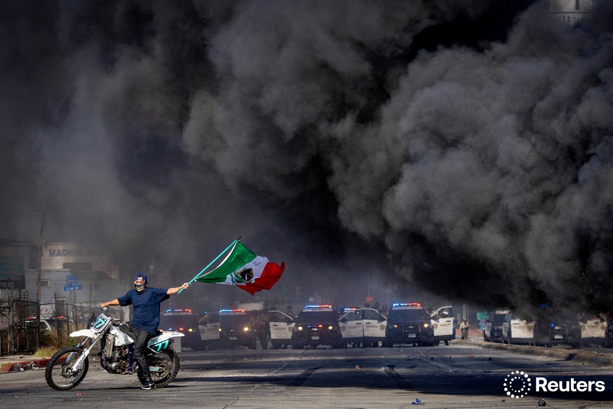 A man on a motorcycle waves a Mexican flag as smoke rises from a burning car on Atlantic Boulevard, during a standoff by protesters and law enforcement, following multiple detentions by ICE in the Los Angeles County city of Compton, California. Photo by <a href="/Photospice/">barbaradavidson</a>