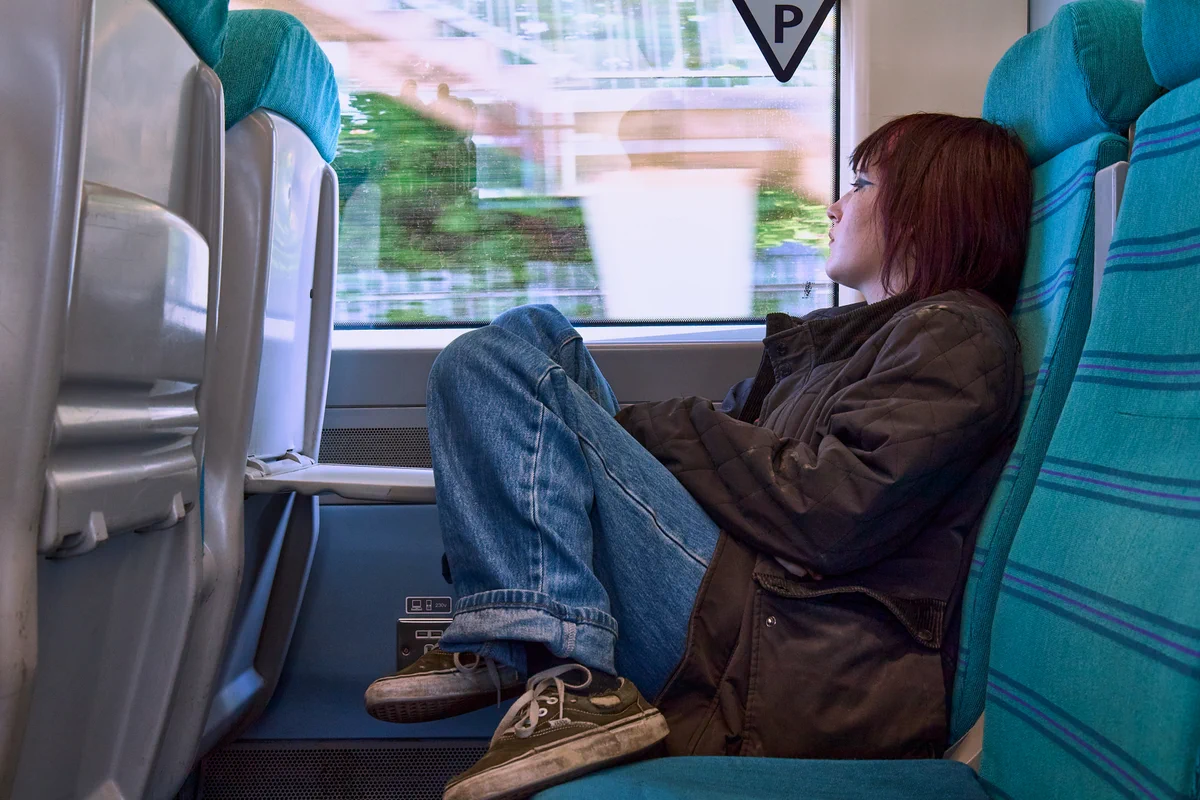 Inside a train, a person with reddish-brown hair, wearing a dark jacket and jeans, sits with knees drawn up on a blue seat, gazing out at the fast-moving, blurred view outside the window.