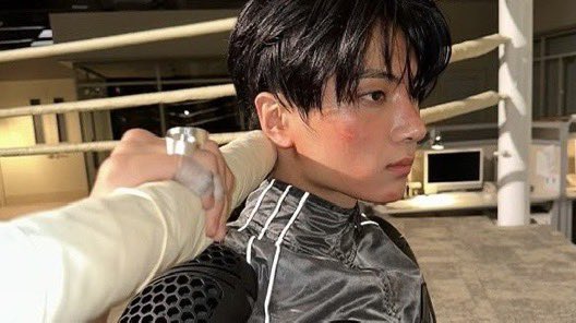 stands in what appears to be a boxing ring, exuding a blend of determination and intensity. The person is dressed in a sleek, silver-gray protective jacket adorned with reflective stripes and black padded armor, designed for safety and style during combat sports.