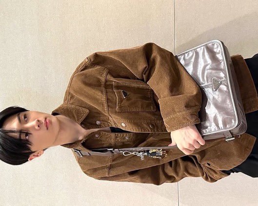 this Prada bag hangs gracefully from the shoulder, with a glossy finish and iconic triangle logo for a subtle nod to high fashion. The bag's sleek design harmonizes with the earthy tones of the jacket, and the bag strap is adorned with a unique keychain featuring a small figure.