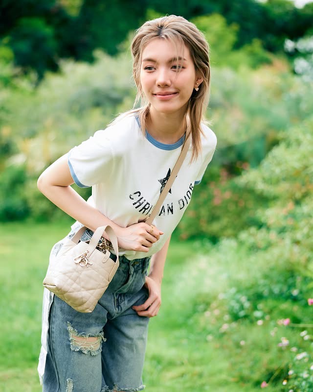 สาวสวยที่มาเติมเต็มความสดใสคือ ออม กรณ์นภัส เธอมาในเสื้อยืดคอกลมสีขาวพิมพ์ลาย "CHRISTIAN DIOR" โดดเด่นด้วยดาวห้าแฉกสีน้ำเงิน จับคู่กับกางเกงยีนส์ฟอกสีอ่อนที่มีดีเทลขาดเล็กน้อย ลุคนี้ตอกย้ำความเป็นสาวสมัยใหม่ที่รักความสบายแต่ไม่ทิ้งสไตล์ สิ่งที่เป็นไฮไลต์คือกระเป๋าถือใบเล็กทรง