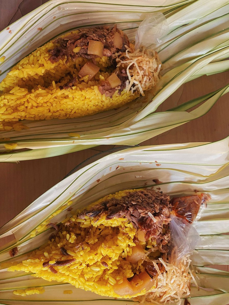 Dua porsi nasi kuning Manado yang disajikan di atas daun woka, lengkap dengan semur daging, kentang, kering kentang, abon, dan sambal dalam plastik kecil. 