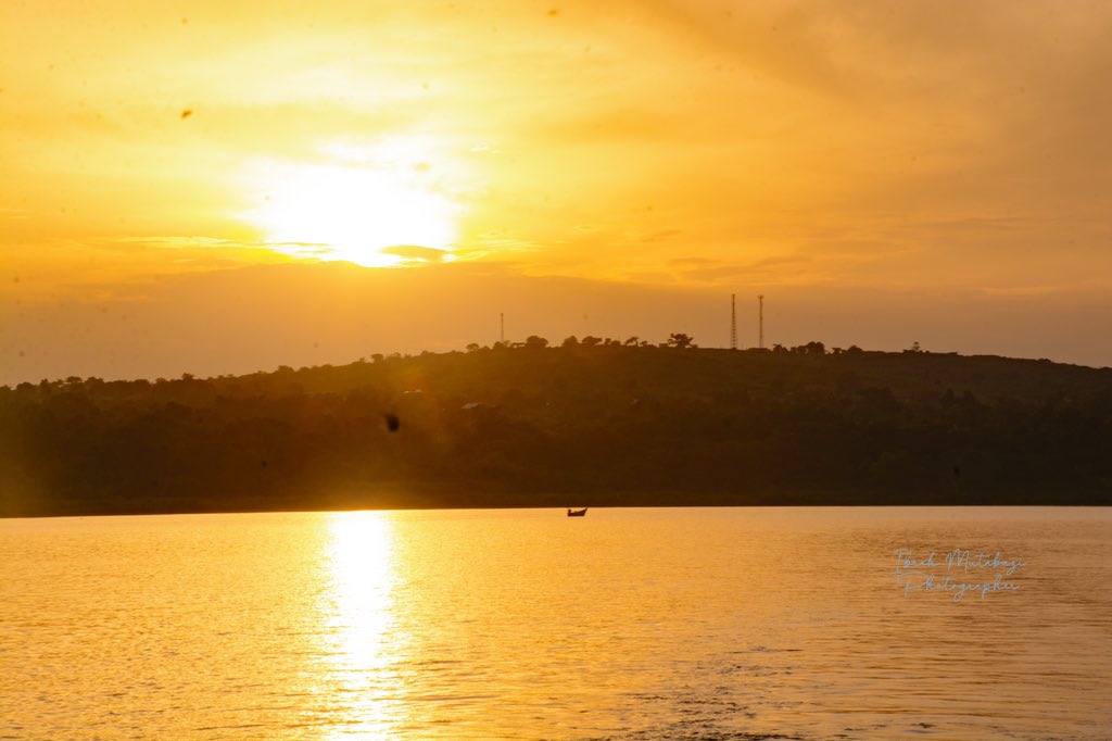 Golden Tranquility – A lone boat sails gently across the River Nile as the sun sets behind the hills of Jinja, Uganda, casting a breathtaking golden glow over the water.