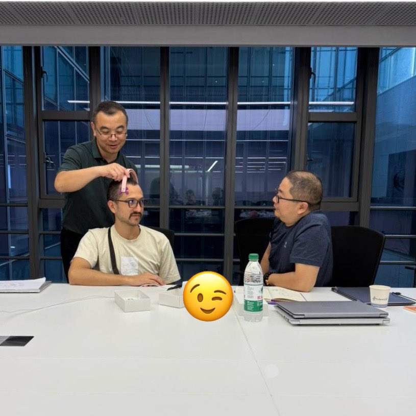 Three men are gathered in a design meeting inside a modern glass-walled conference room. One man is seated, wearing a light-colored t-shirt and glasses, while another stands behind him, holding a pink prototype or measuring tool to his forehead. Across the table, a third man listens and observes closely. The table holds open product boxes, a water bottle, notebooks, and laptops. A winking emoji partially covers the foreground. The scene suggests collaborative evaluation of a wearable device concept.