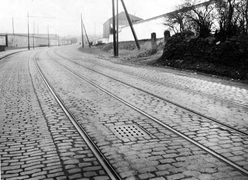 The approach to Elland Road from under the railway bridge. Leeds Greyhound Stadium on the right, on the left  what at the time was the Leeds Speedway on Fullerton Park with the Leeds United ground just visible. One of the stone gateposts on the right still stands today, 1930.