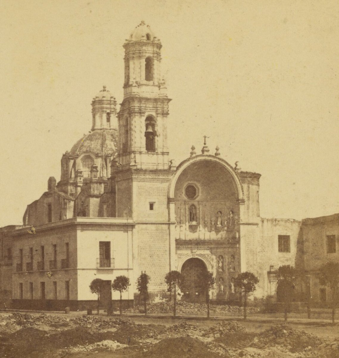 El templo de San Juan de Dios fotografiado alrededor de 1863-1870 por el francés Julio Michaud. Lo impresionante es el entorno que rodea el templo, pues se encuentra al norte de la Alameda central. Noten los montículos de tierra, así como los pequeños árboles. También es
