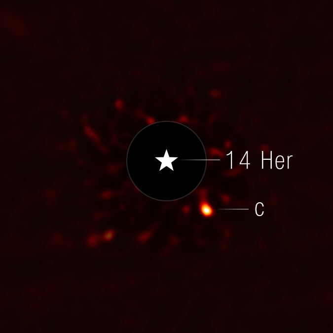 This image shows the exoplanet 14 Herculis c. The view is mostly black, with very faint red splotches in the central region of the image. At the center of the image, there is a black circle, and in the center of that, there is a star symbol representing a real star. This black circle blocks the light from the host star. To the lower right of the circle is a fuzzy bright orange circle, which is the exoplanet.