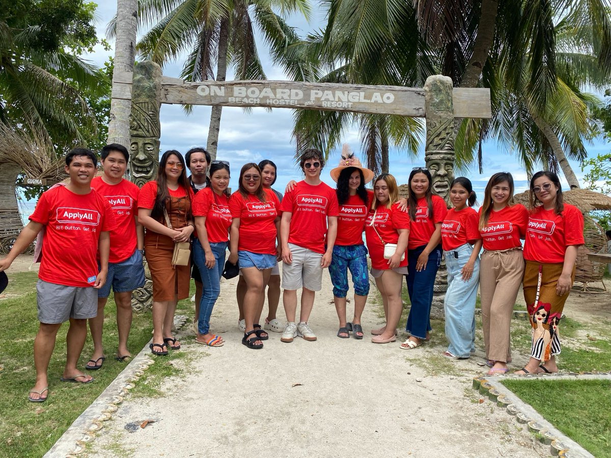 A dozen people lined up for a team photo at a beach resort. Everyone on the team is wearing a redshirt that says "ApplyAll - push button. get job."