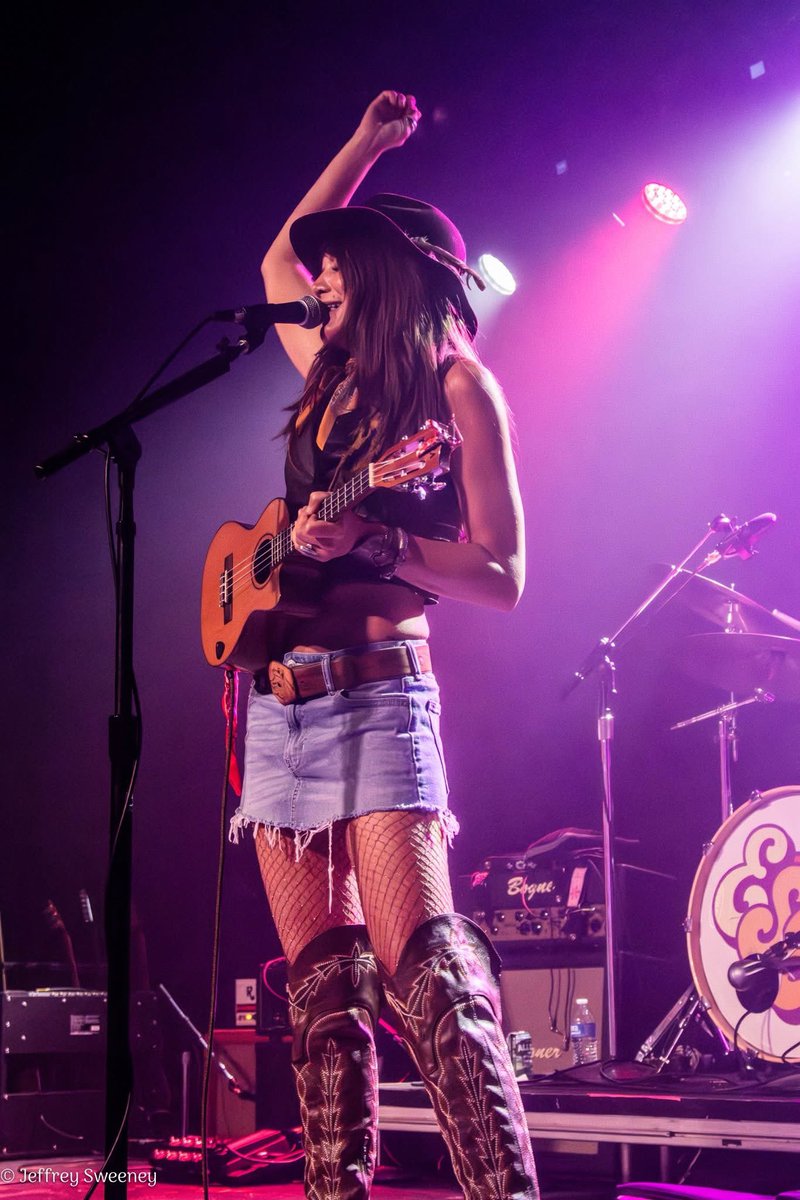 “a dynamic moment during a live musical performance on stage—likely the frontwoman of a band—is mid-song with one arm raised triumphantly while singing into a mic. She’s playing a ukulele and radiates confidence and charisma”#RaelynNelsonBand