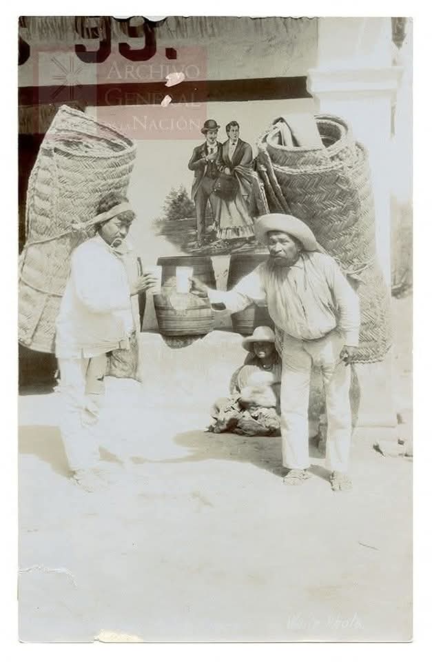 Dos hombres hacen una pausa para tomar pulque, alivio sagrado entre la venta de petates.
La sed y la necesidad no dan tregua.
Detrás, una mujer y su hijo observan en silencio.
México, sin fecha, pero eterno.
📷 Archivo General de la Nación.