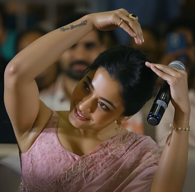 The image features actress Rashmika Mandanna, who is known for her glamorous appearances. She is dressed in a pink saree with intricate detailing, holding a microphone in one hand while posing with her other hand raised above her head. The setting appears to be a public event or a gathering, with an audience visible in the background. The post text, "She never FAILS 🤤", suggests admiration for her consistent style and presence. Rashmika's confident and engaging pose, along with her elegant attire, highlights her status in the fashion and beauty industry.