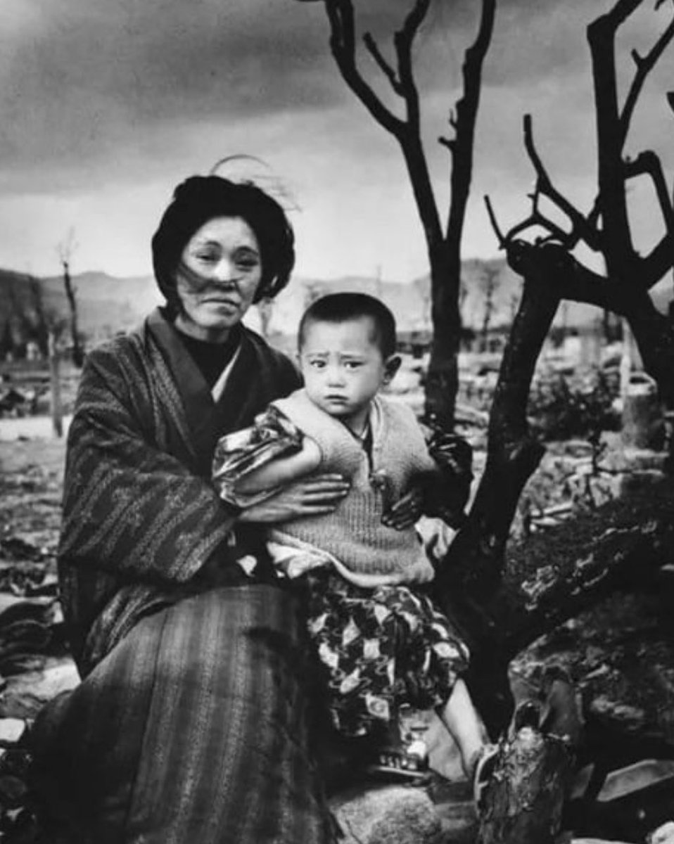 Four months after the atomic bomb was dropped, a Japanese mother and child, dressed in traditional clothing, sit amid the ruins and charred trees of Hiroshima. December 1945.
