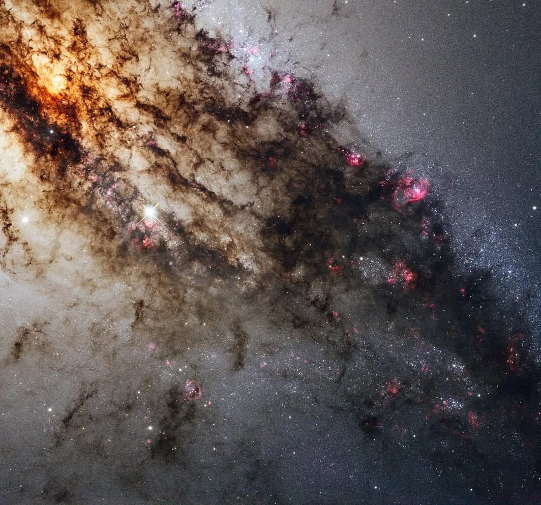 Cosmology0's tweet image. Centaurus A, features a bright orange region in its upper left, with dark dust flowing diagonally downwards. The background is a hazy space filled with distant stars, and pink areas of new star formation are visible within the dust.