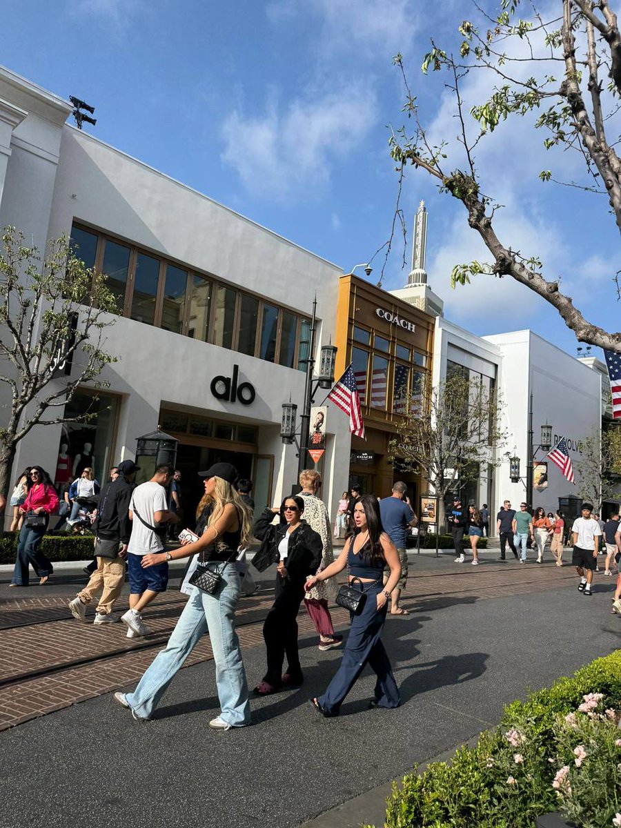 Busy shopping street with stores like Alo and Coach, decorated with American flags, on a sunny day in June 2025.