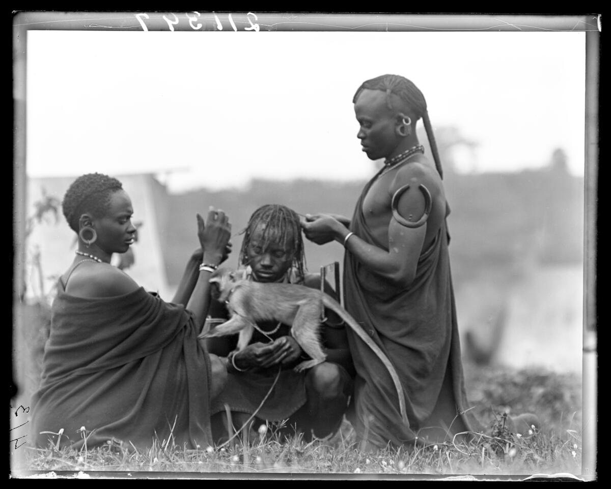 Kikuyu women 
Braiding man's hair while he holds monkey J.T. Junior, Kenya, 1909-1911