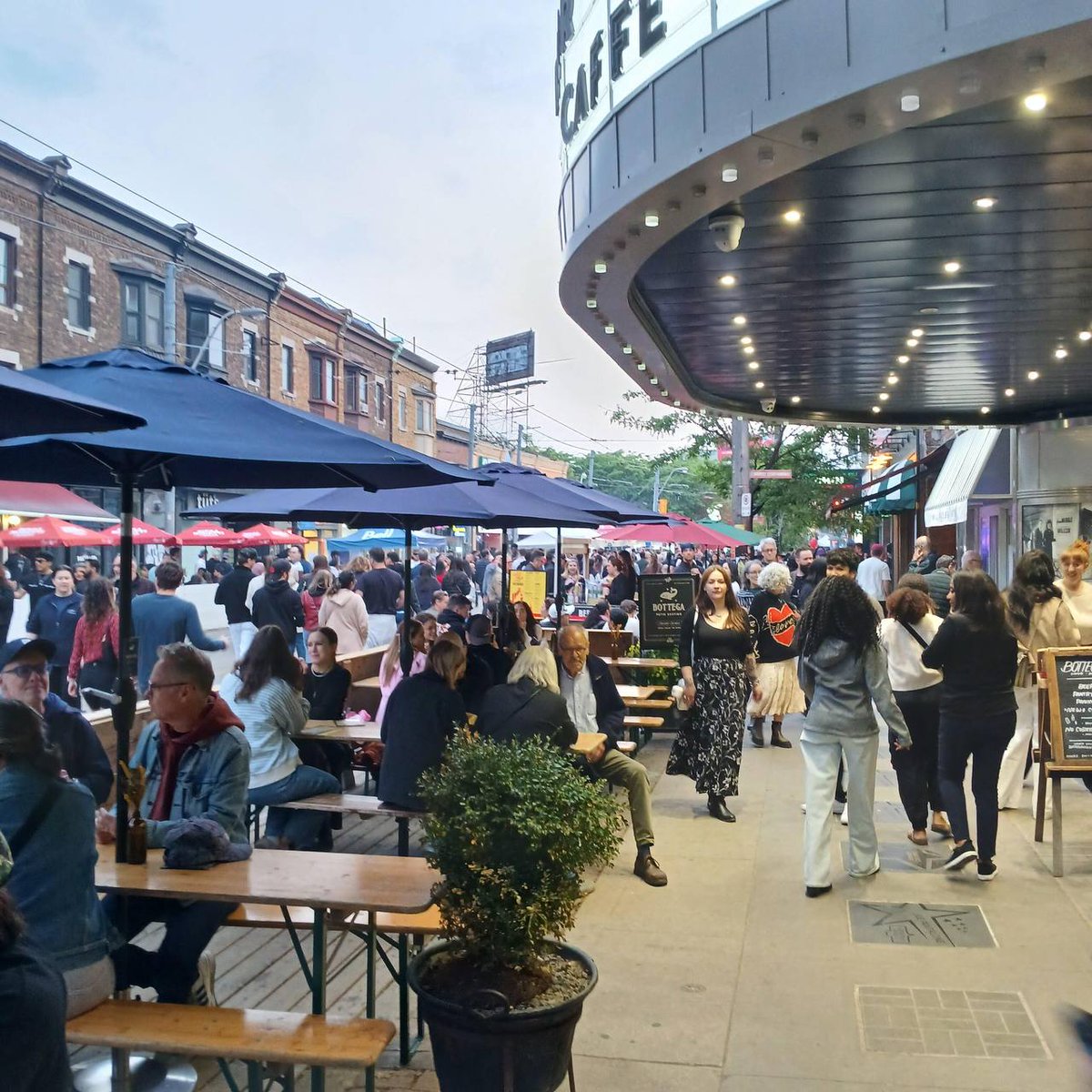 Busy outdoor café scene with people dining at wooden tables under colorful umbrellas next to a theater with a "CAFE" sign on a lively street with shops and pedestrians.